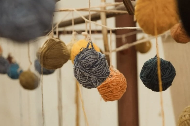 Multiple balls of yarn in assorted colors hanging from a wooden rack against a light neutral wall.