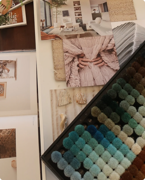 A styled interior design display featuring a large black tray filled with textured pom-pom fabric swatches in a gradient of blue, green, beige, and brown, alongside printed room inspiration photos and fabric swatches.