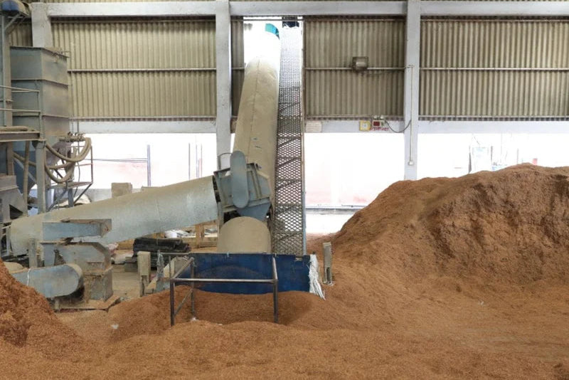 A large industrial silo and conveyor system processing a bulk pile of brown wood chips or mulch inside a warehouse.