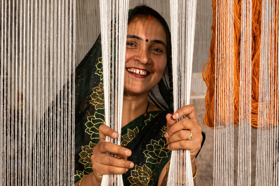 A smiling woman in a dark green embroidered sari holds white hanging threads in front of her face, with orange yarn visible on the right side.