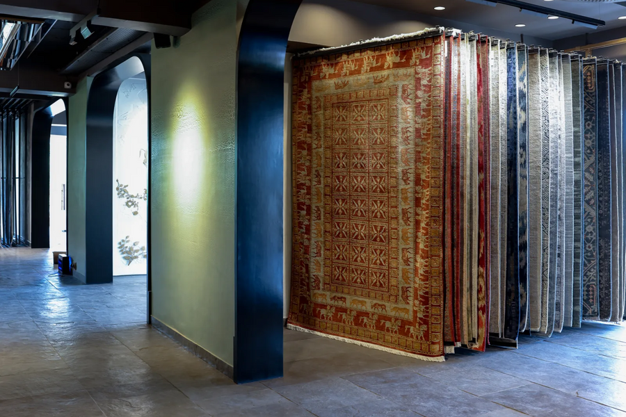 A large patterned area rug hangs in a showroom next to a wall of hanging rugs in various colors and patterns, with a modern interior featuring concrete floors and dark columns.