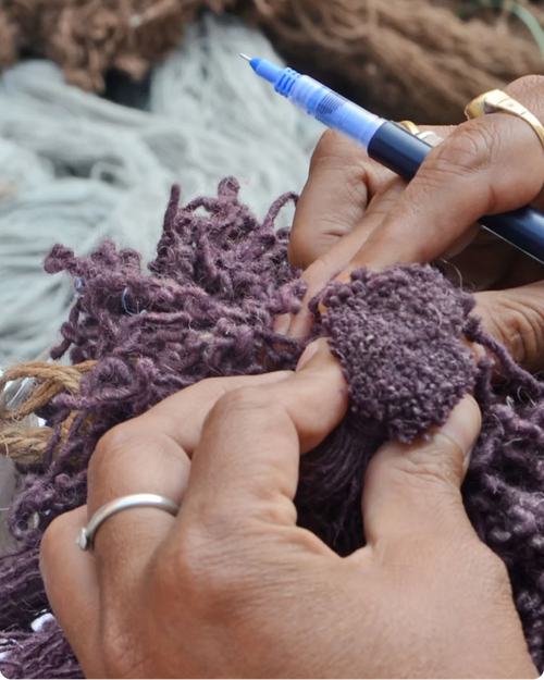 Close-up of hands knitting with a blue-handled needle and dark purple yarn, with rings on the fingers and a soft-focus background of more yarn.