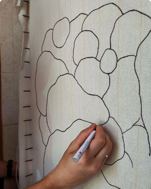 A hand holding a white marker draws a floral pattern outline on a white textured fabric panel.