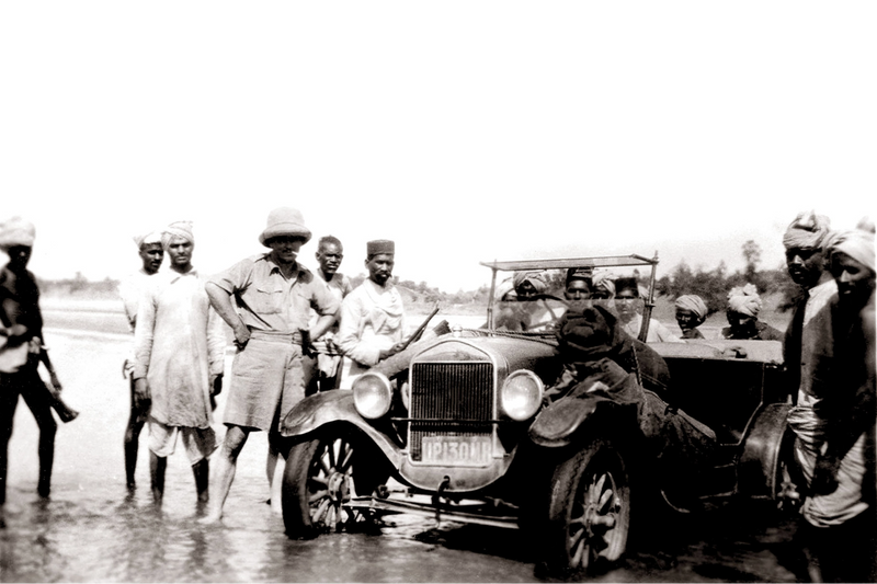Black and white historical photograph showing a vintage open-top car on a road with several men standing and sitting around it.