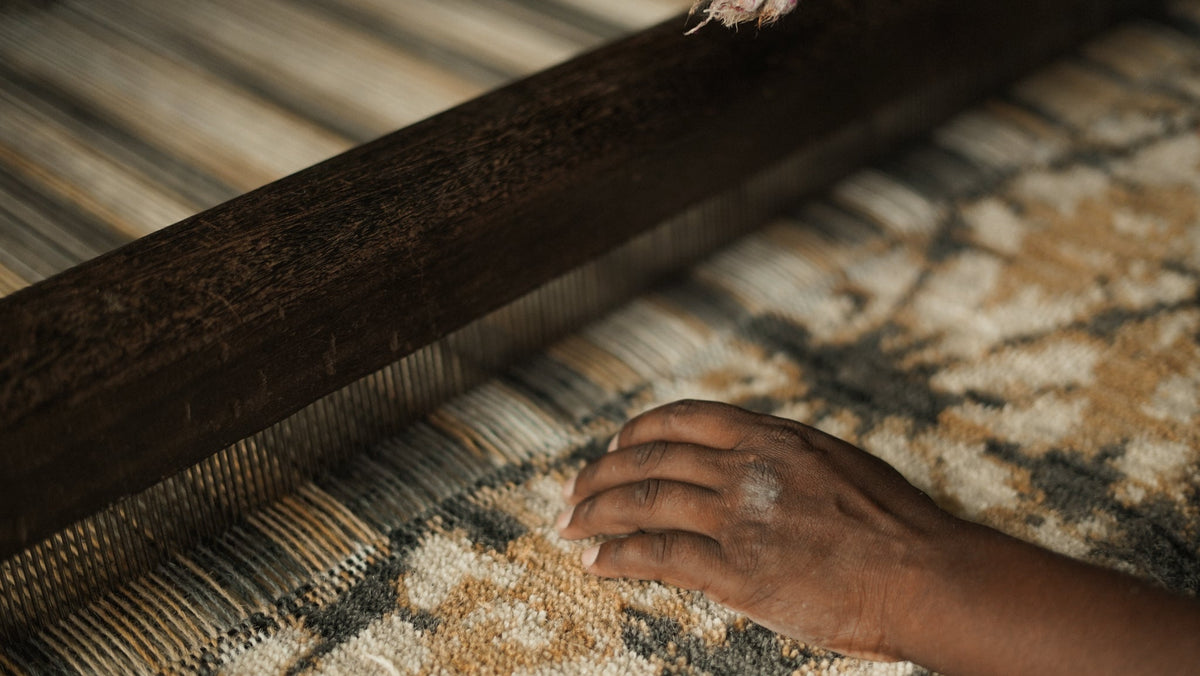 A close-up of a hand weaving a patterned rug with a dark wooden loom in the foreground.