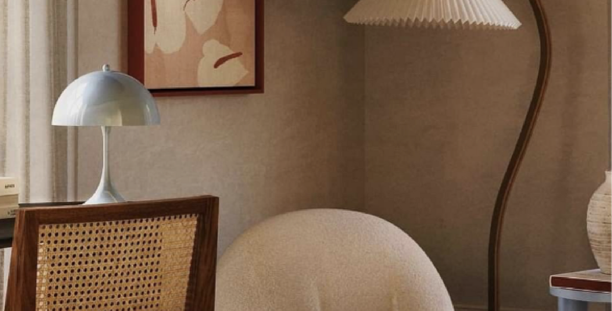A styled interior corner featuring a white dome-shaped table lamp on a wooden chair back, a cream upholstered chair, a pleated floor lamp, and a framed abstract print on a textured beige wall.