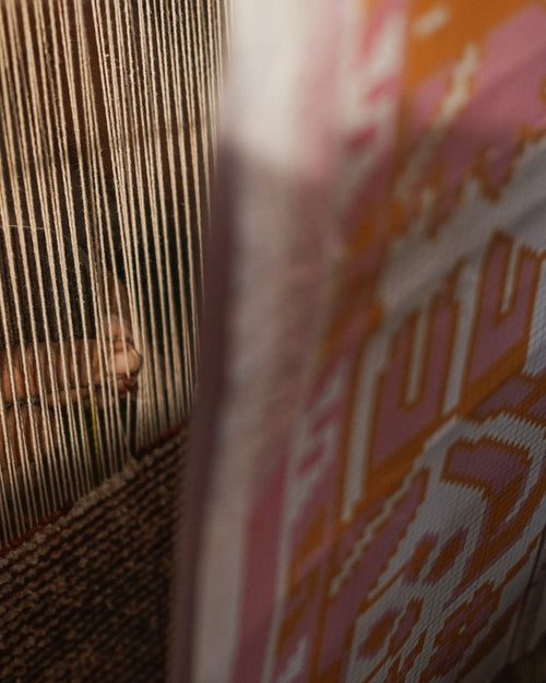 A close-up view of a woven textile with vertical beige and brown stripes on the left and a pink, orange, and white patterned section on the right, partially blurred in the foreground.